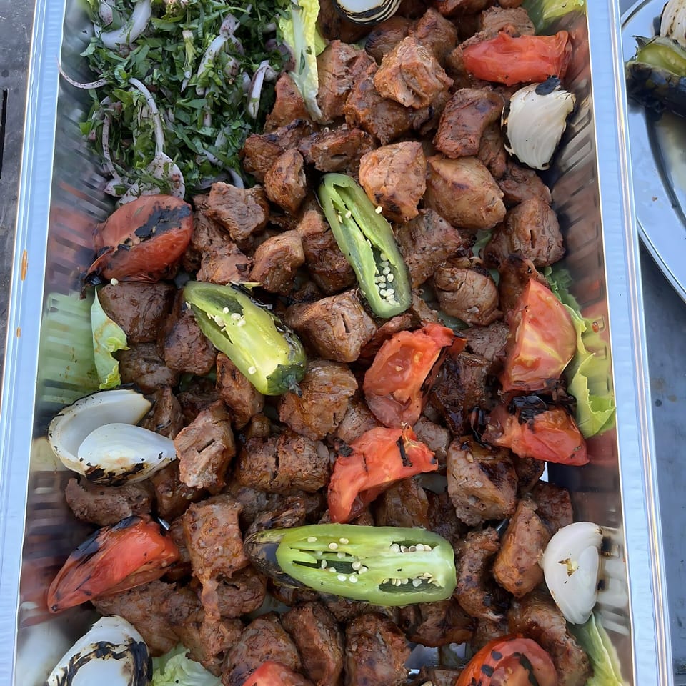 Grilled Mediterranean meat and vegetables, including tomatoes, peppers, and onions, in a metal tray.