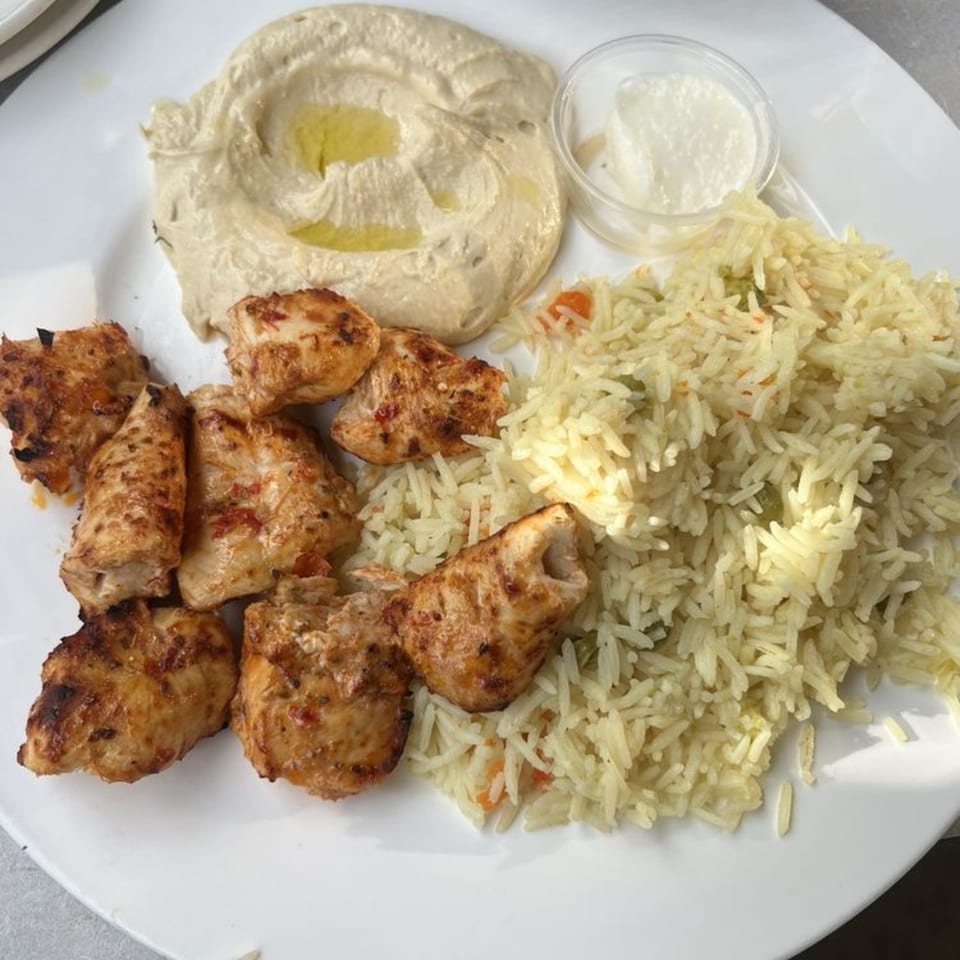 Grilled chicken pieces, rice, hummus, and a small dish of white sauce on a white plate.