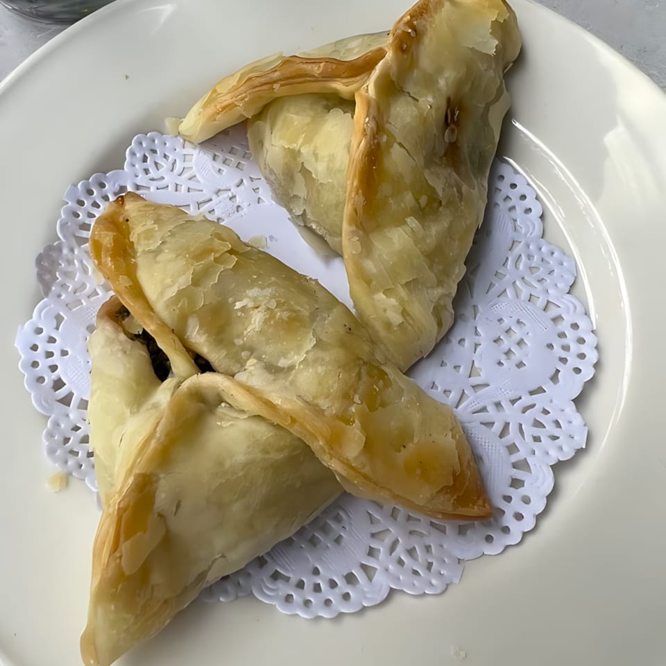Three golden-brown, triangular Mediterranean pastries, likely fatayer or sambousek, on a white plate with a doily.