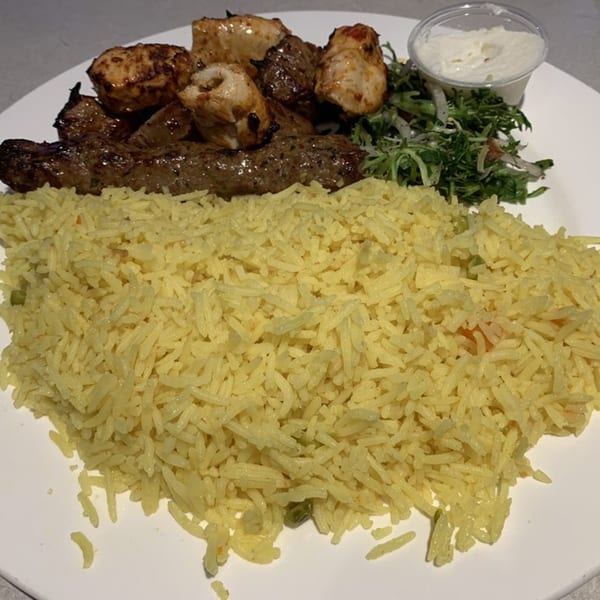 A plate of Mediterranean food with yellow rice, grilled chicken and beef skewers, salad, and a side of white sauce.